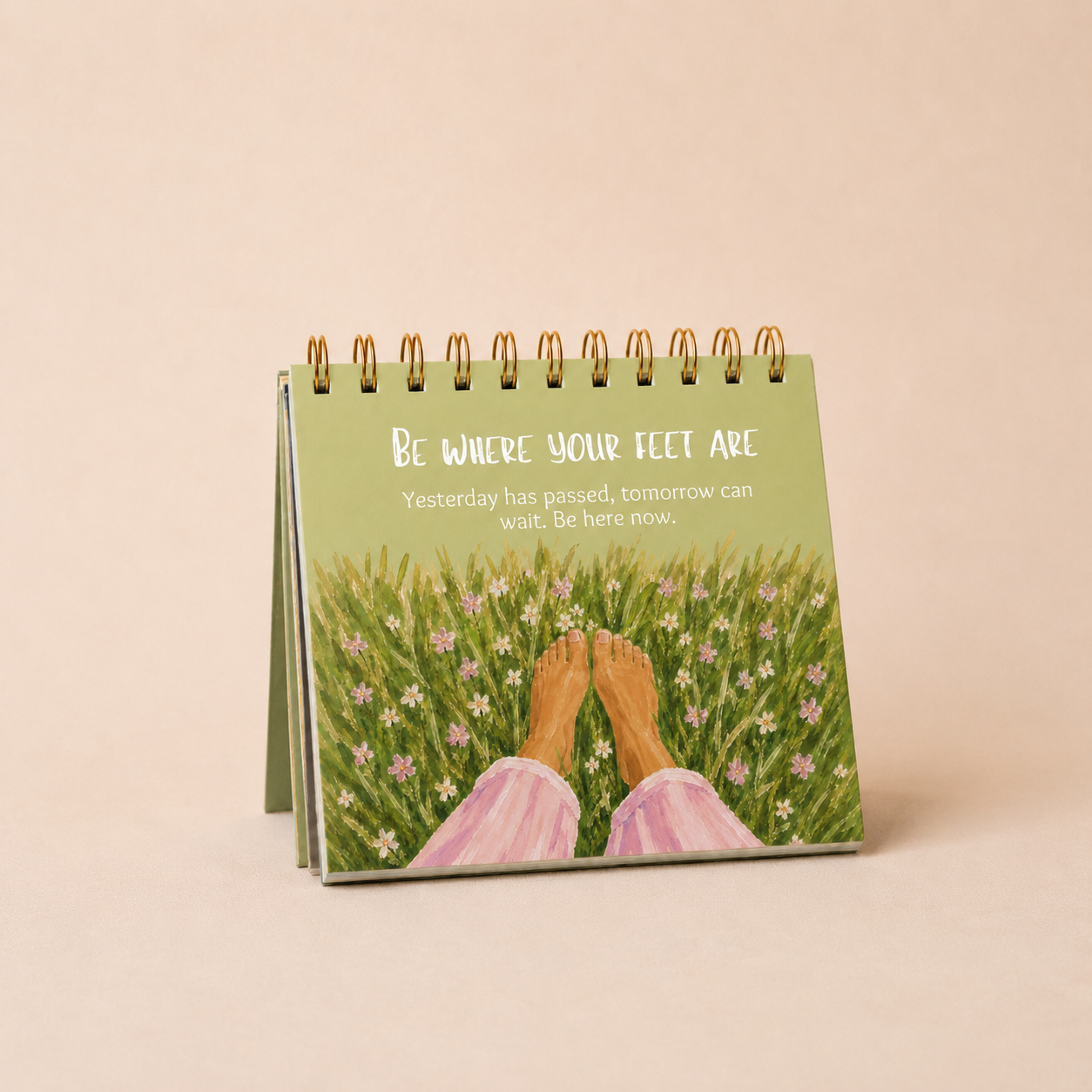 A spiral-bound desk calendar or art print standing upright, showing a painted scene of bare feet in a grassy field with small pink and white flowers. White text on the cover reads, 'Be where your feet are.