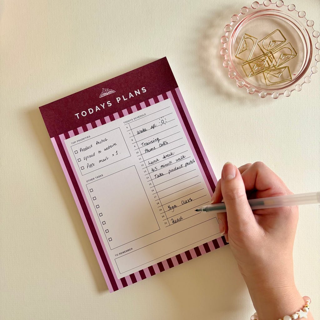 A hand holding a pen writes on a 'TODAYS PLANS' notepad, placed on a light beige surface next to a pink glass dish with gold paperclips.