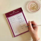 A hand holding a pen writes on a 'TODAYS PLANS' notepad, placed on a light beige surface next to a pink glass dish with gold paperclips.