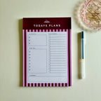 A 'TODAYS PLANS' notepad with a dark red header and pink-and-white striped borders, shown on a light beige surface with a pen and a decorative dish nearby.
