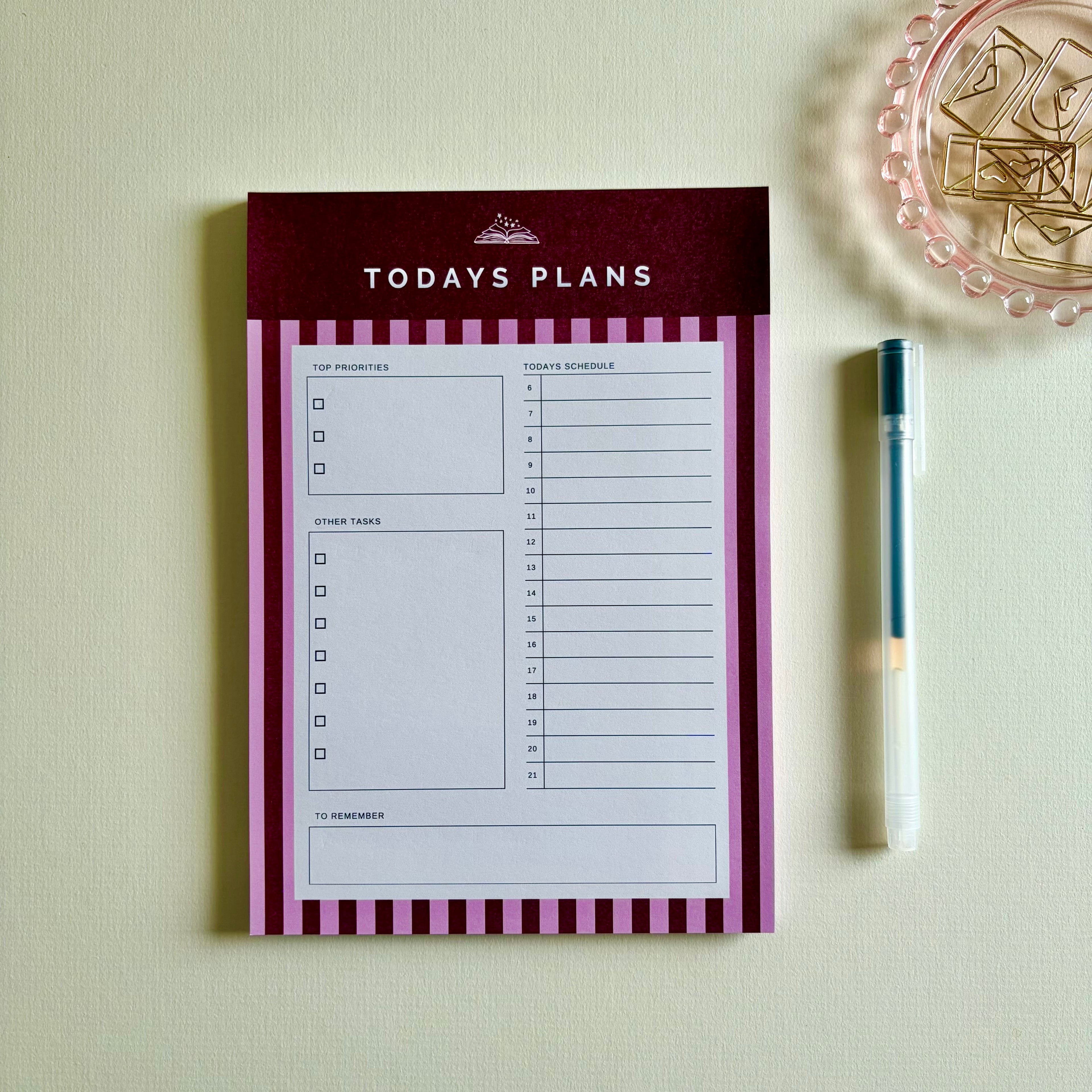 A 'TODAYS PLANS' notepad with a dark red header and pink-and-white striped borders, shown on a light beige surface with a pen and a decorative dish nearby.