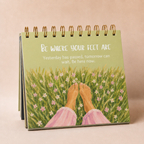 A spiral-bound desk calendar or art print standing upright, showing a painted scene of bare feet in a grassy field with small pink and white flowers. White text on the cover reads, 'Be where your feet are.