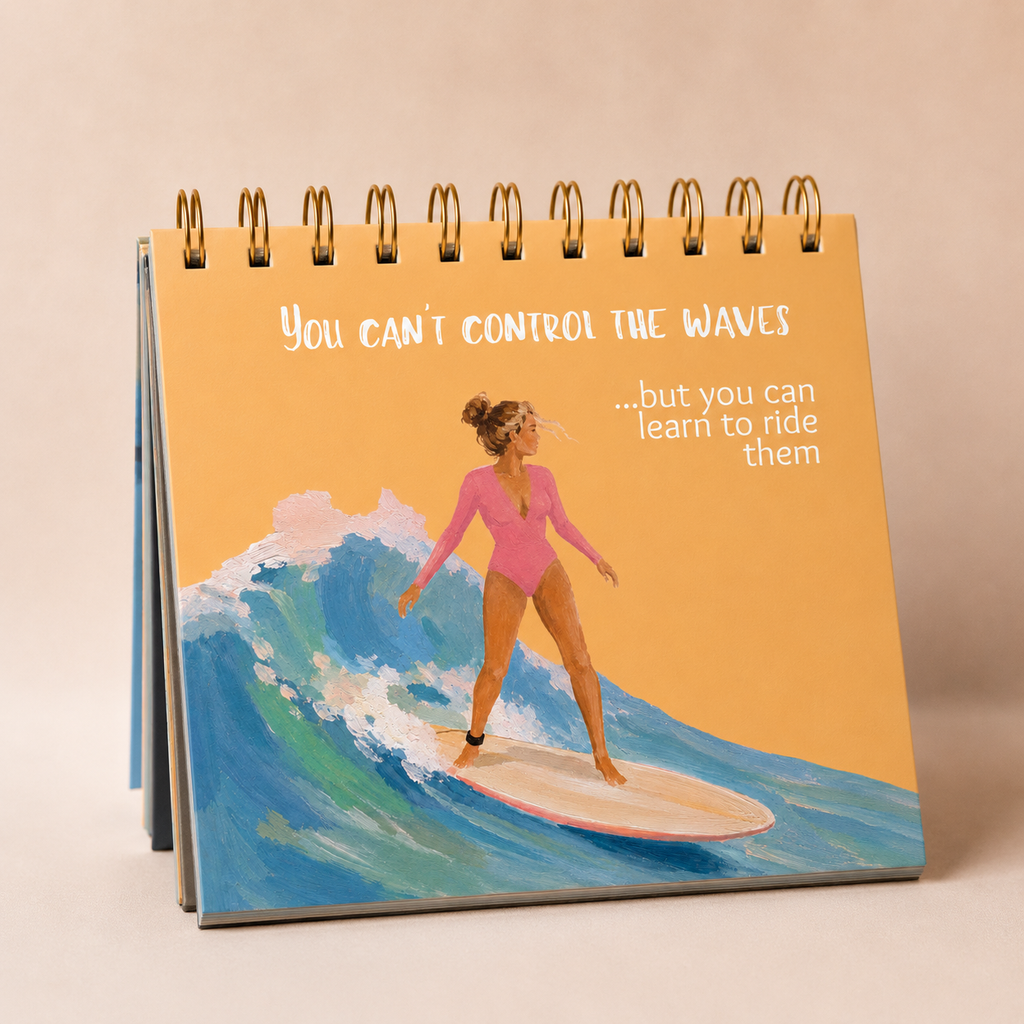 A spiral-bound desk flip book or notebook standing upright, showing an illustration of a woman in a pink swimsuit surfing on a blue wave against a warm tan background. White text on the cover reads, 'You can't control the waves .