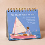 A spiral-bound desk calendar or tabletop print with a blue cover, featuring an illustration of a woman in a pink sailboat on the water and the text, 'YOU HAVEN'T MISSED THE BOAT.'.